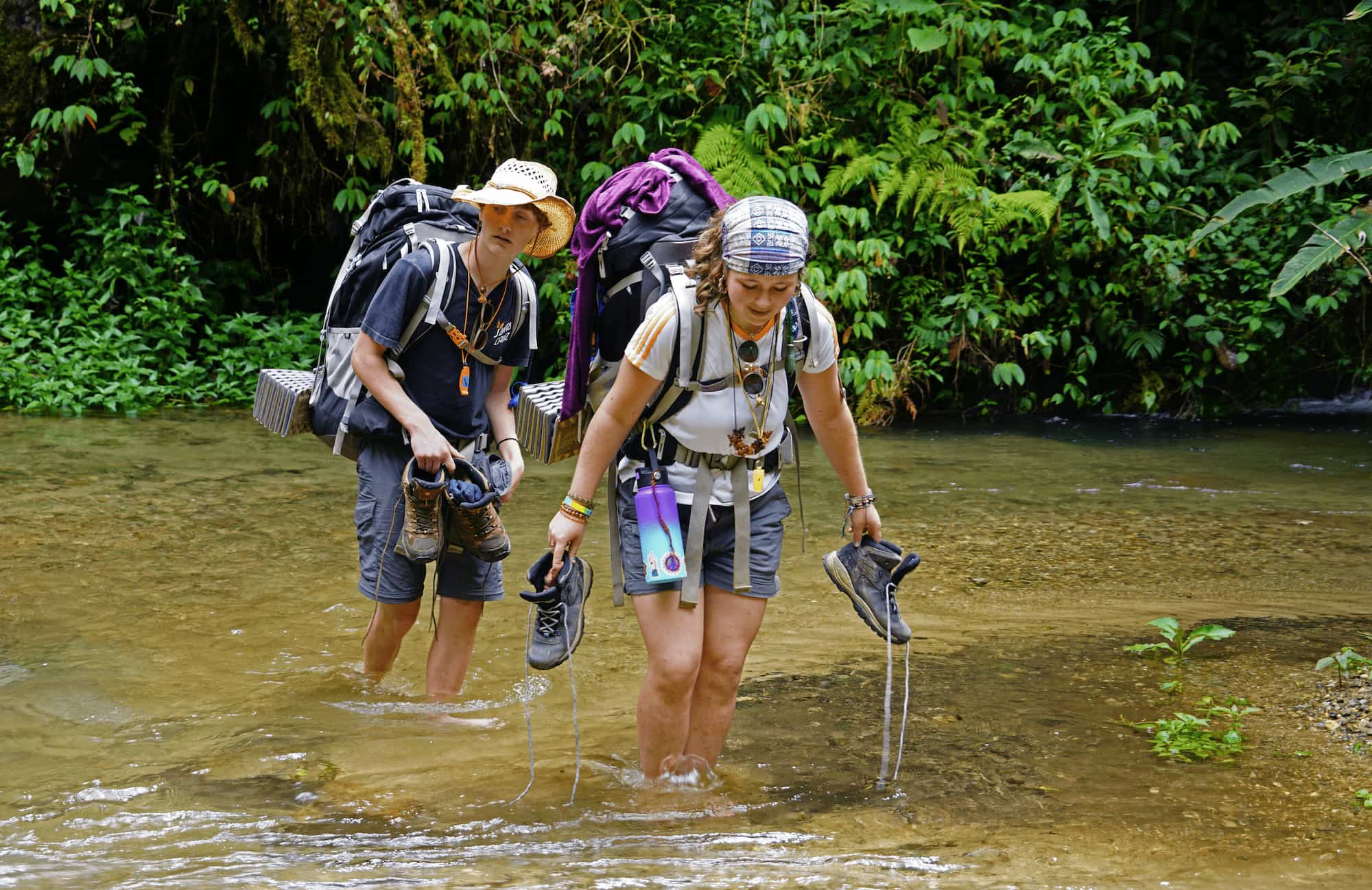 Two people crossing a river by foot, carrying hiking gear