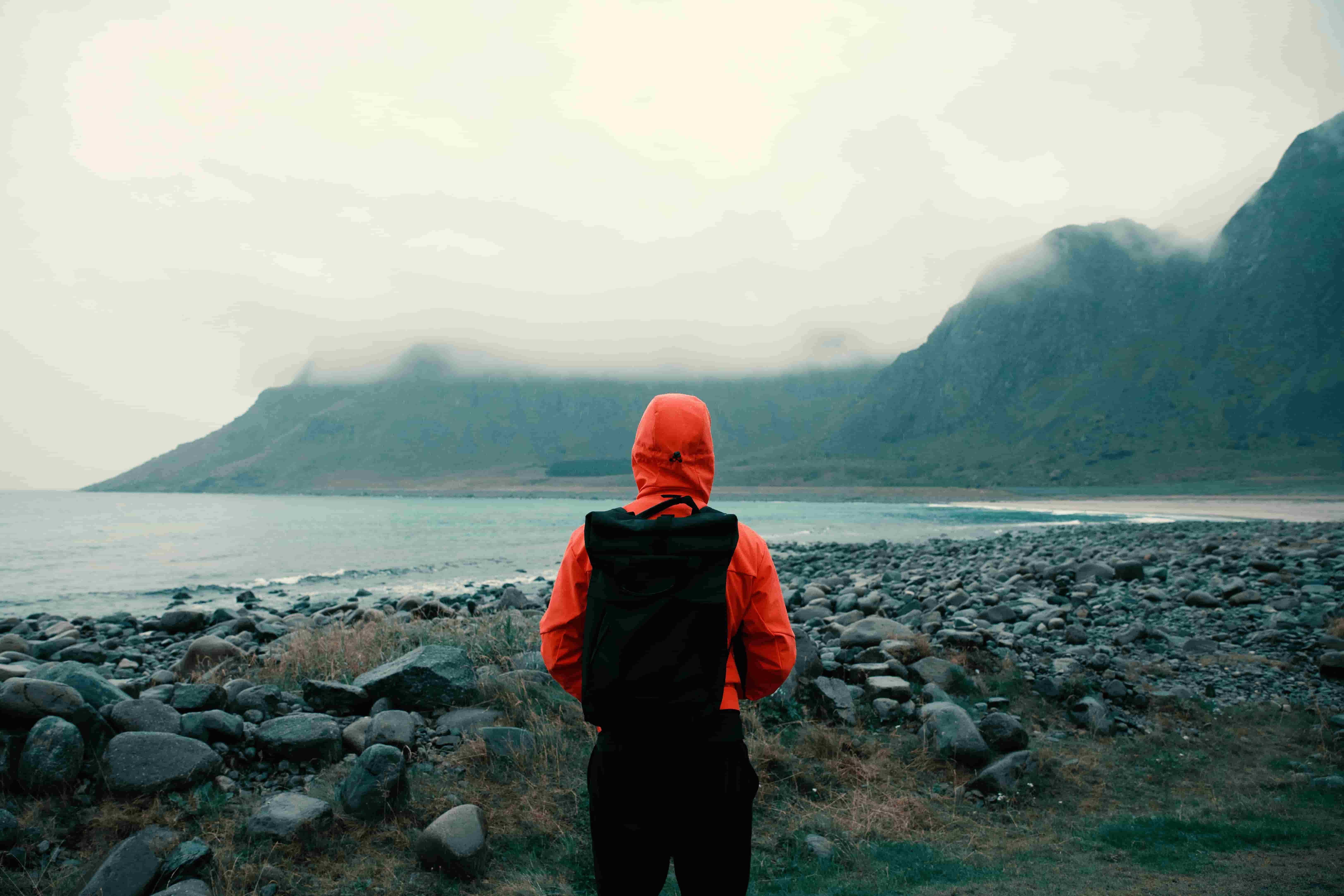 A man in a red rainjacket looking over water towards amisty mountain